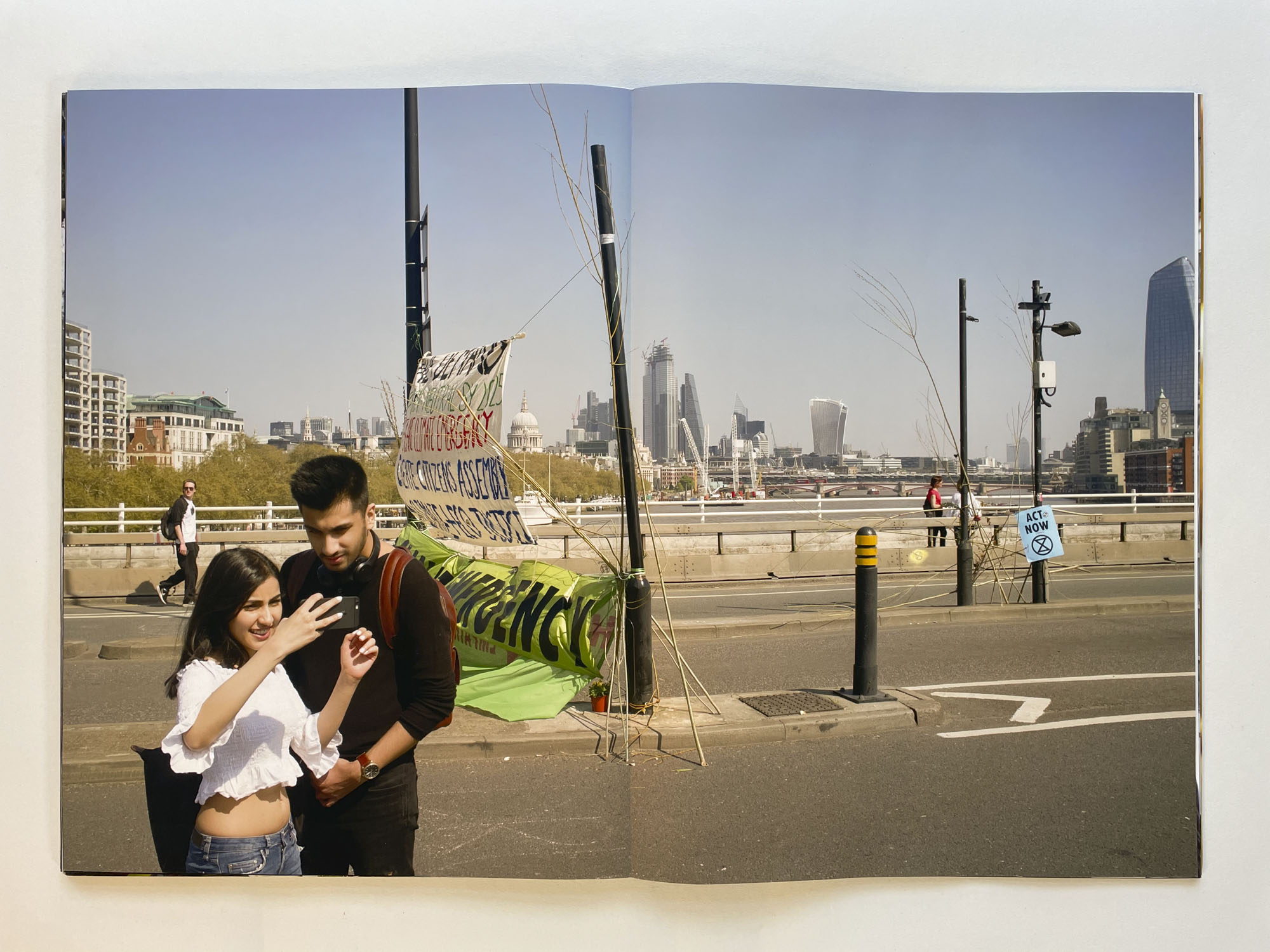 a young woman takes a selfie with her boyfriend in front of a nearly deserted bridge with a climate protest banner visible behind them