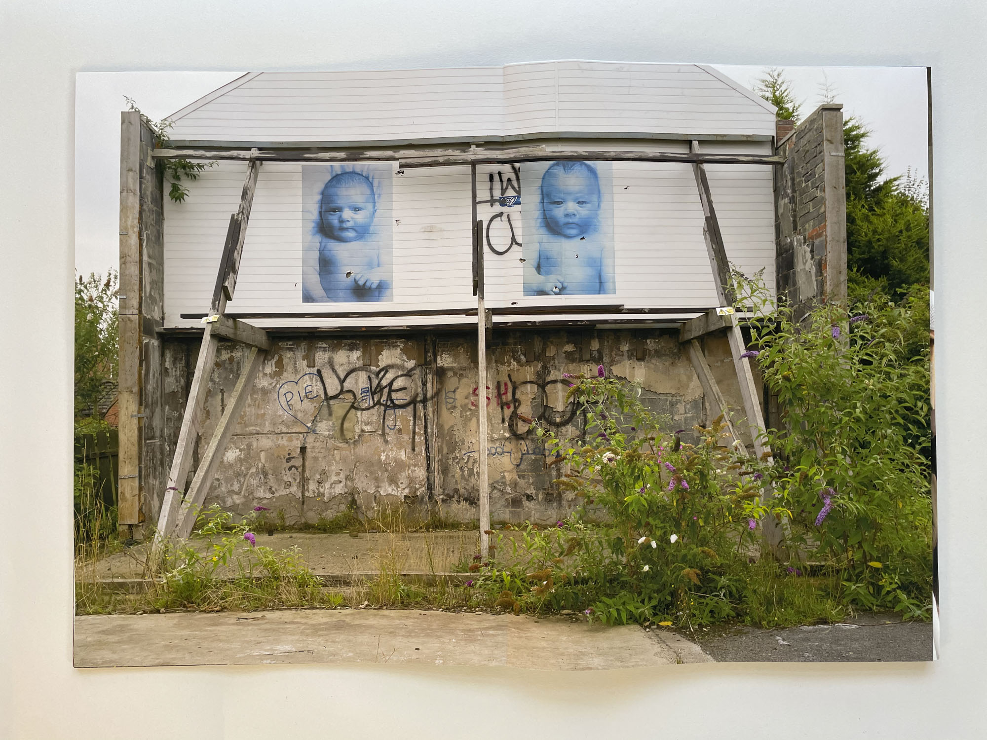 the blank end of a house is buttressed by wooden supports, on the wall are two large blue photographic prints of babies